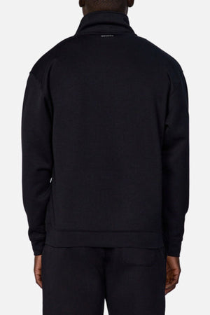 Seen from the back, a person wears the Industrie The Tech Staunton Track Top and matching pants in black. The athletic jacket has a high collar and a small white logo at the top, set against a plain light background.