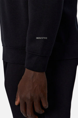 A close-up of a person’s arm and hand wearing the Industrie The Tech Staunton Track Top, featuring "INDUSTRIE" printed in white on the sleeve. The black athletic fabric contrasts with a plain, light background.