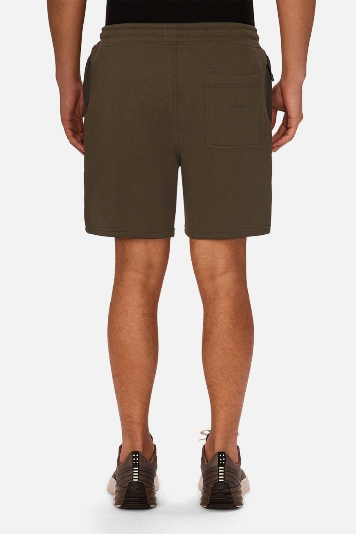 A person is standing facing away, wearing olive green Industrie The Tech Track Short, a black top, and gray sneakers. The athletic shorts feature a back pocket on the right side. The background is plain white.