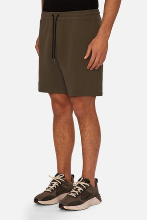 A person wearing Industrie The Tech Track Short in olive green, paired with a black top and brown athletic shoes with white soles, stands against a plain white background.