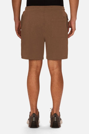 A person is seen from behind wearing the Industrie The Tech Track Short in brown, featuring a drawstring waist, along with black sneakers, standing against a plain white background.