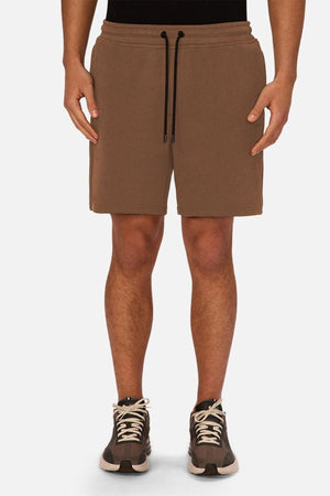 A person wearing the Industrie The Tech Track Short in brown with a drawstring waist, paired with brown and black sneakers featuring white laces and a black shirt, stands against a plain white background—an effortlessly stylish look in men’s activewear.