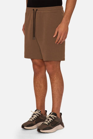 A person is wearing the Industrie The Tech Track Short in brown, paired with a black shirt and brown athletic shoes with white soles, standing against a plain white background.