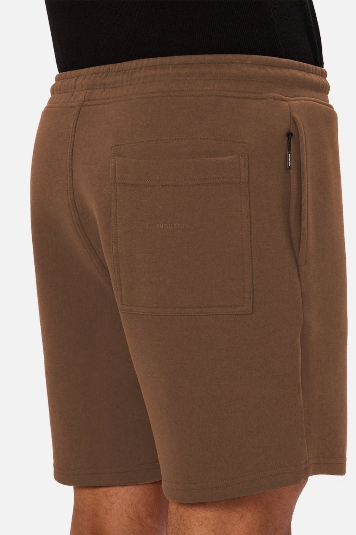 A person wearing the Industrie The Tech Track Short—brown men’s athletic shorts featuring a back pocket, drawstring waist, and a zippered side pocket—standing against a white background.