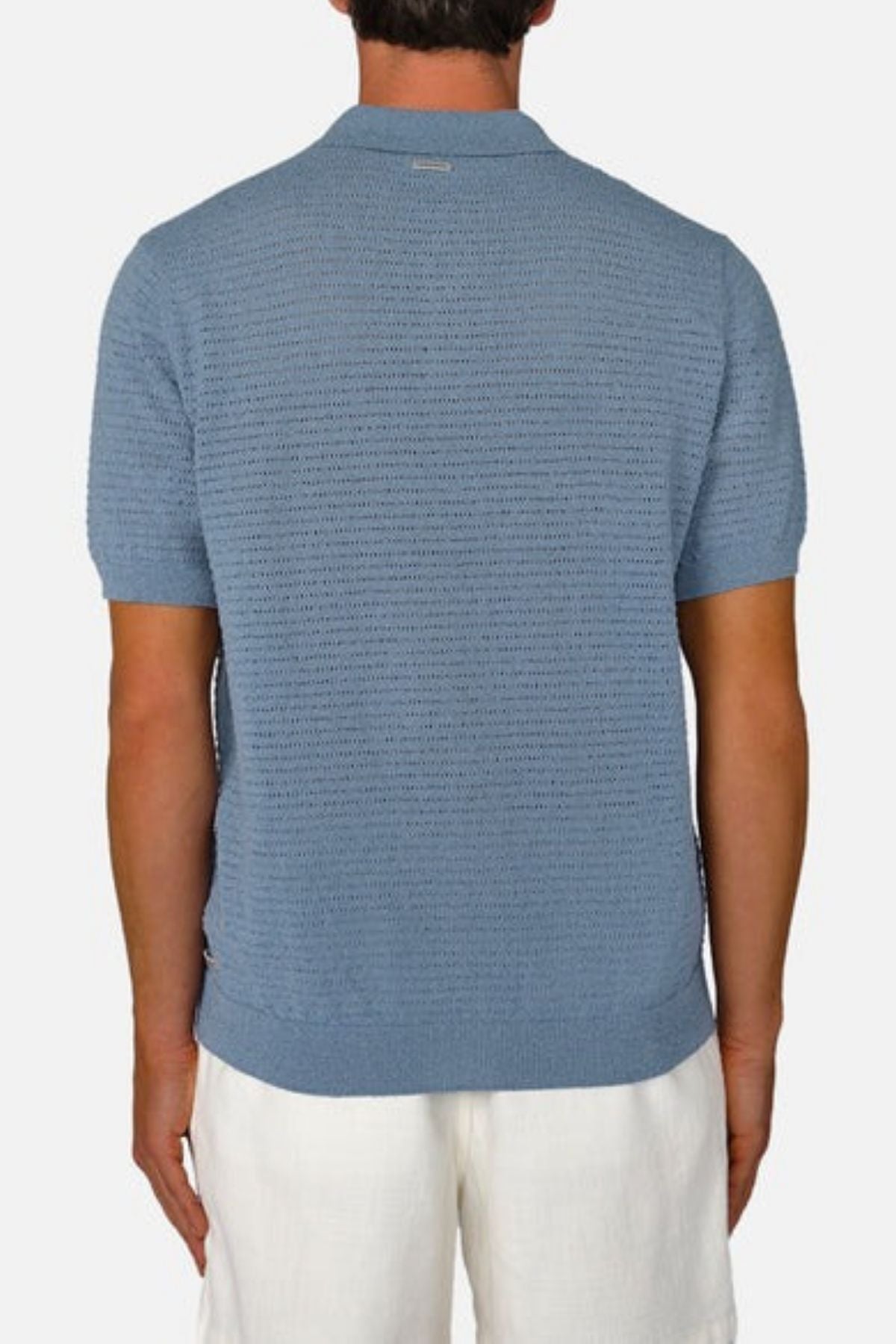 The individual is seen from behind against a simple backdrop, dressed in a light blue short-sleeve Industrie The Terracina Polo, featuring a vertical ribbed texture, paired with white pants.