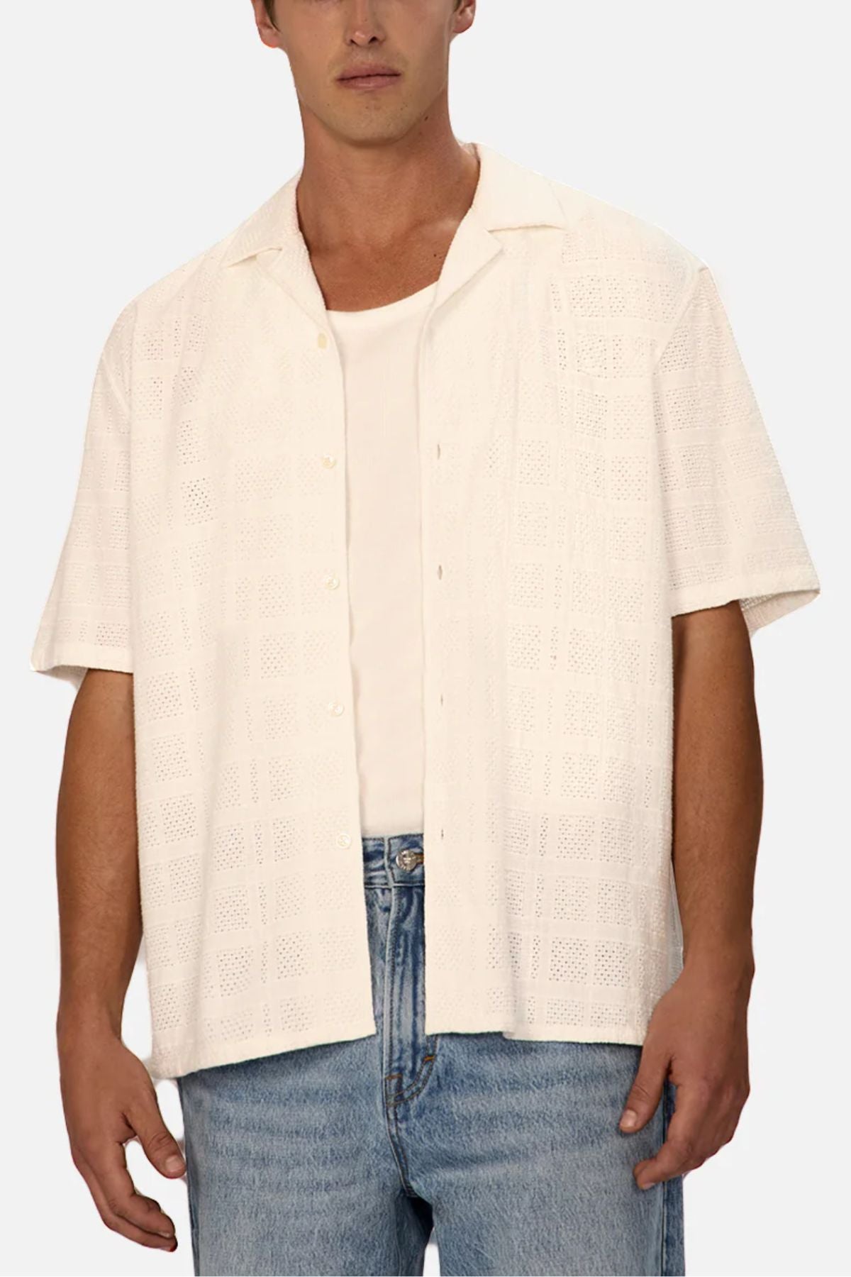 A man wears the Industrie The Venty Short Sleeve Shirt, an off-white, lightweight cotton shirt with a relaxed fit and subtle textured pattern, over a white tank top and light blue jeans against a plain backdrop.
