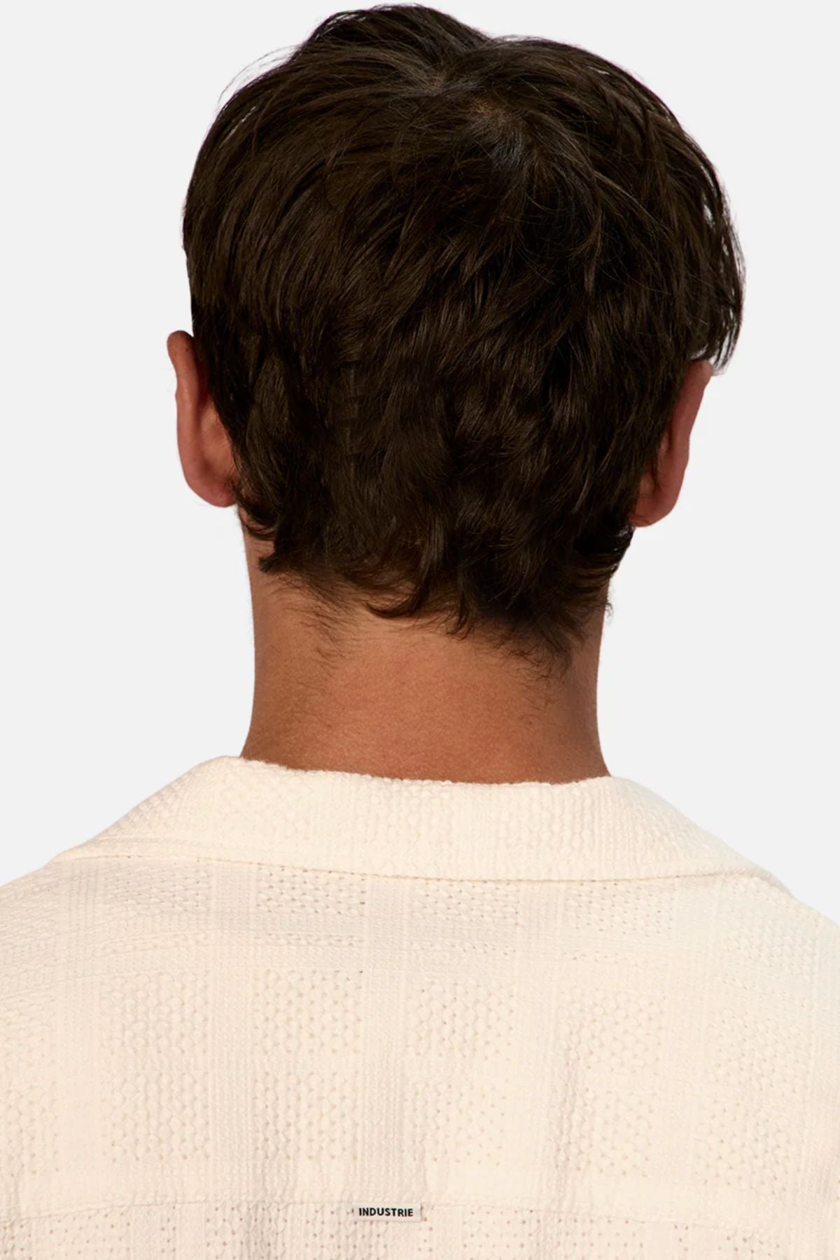 A person with short brown hair is shown from behind, wearing the Industrie The Venty Short Sleeve Shirt in textured white cotton. The background is plain and light-colored.