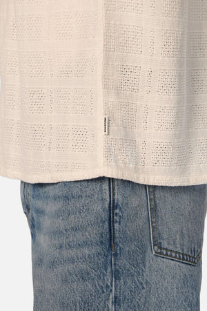 Close-up of a person wearing light blue denim jeans and Industrie The Venty Short Sleeve Shirt—a cream, lightweight cotton shirt with a subtle grid pattern and a small white tag with black text on the side seam.