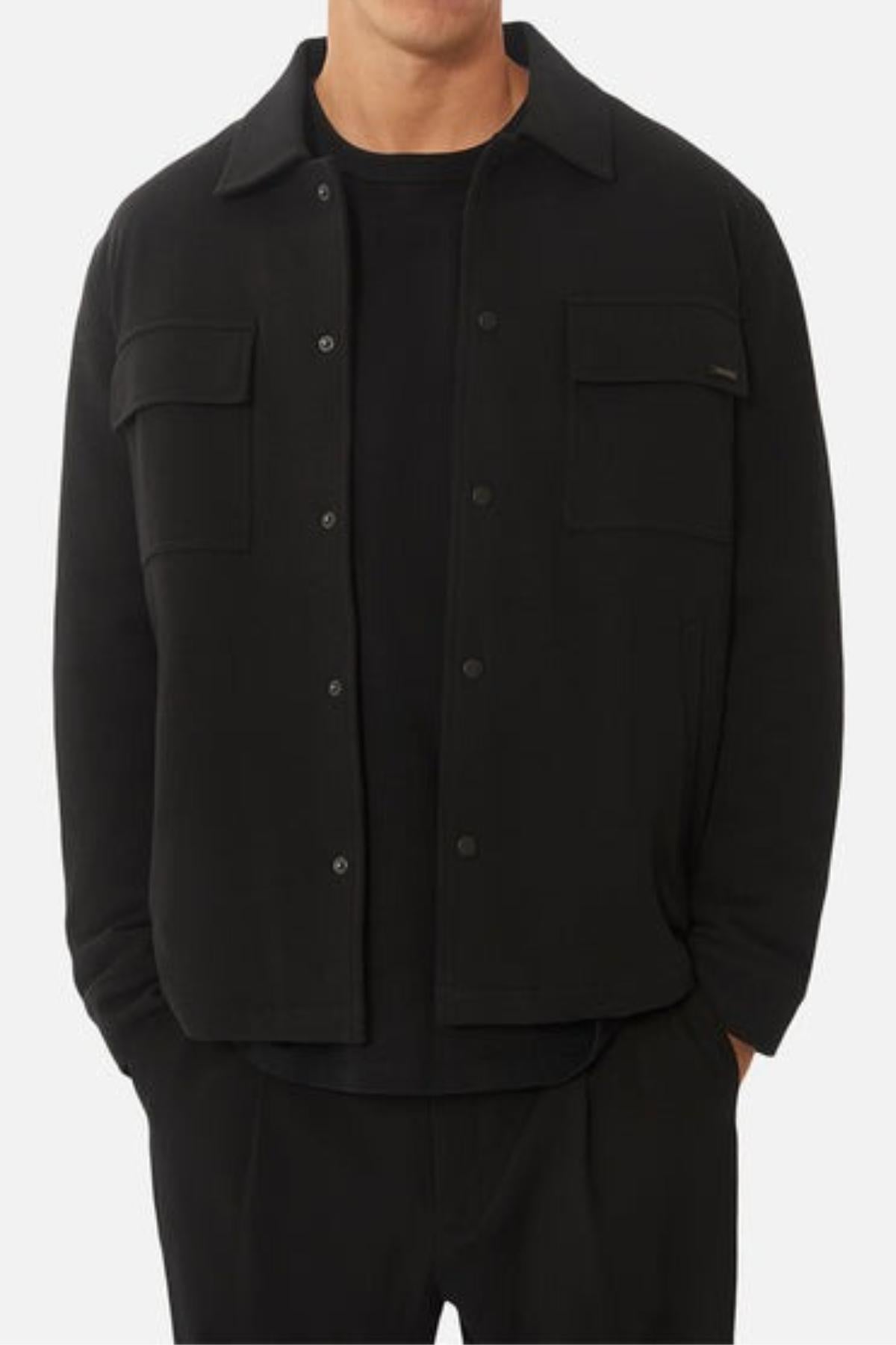 A man stands against a plain background with hands in his pockets, wearing the Industrie The Westport Jacket in black, styled over a black t-shirt and black pants. Twin chest pockets complete the jacket’s look.