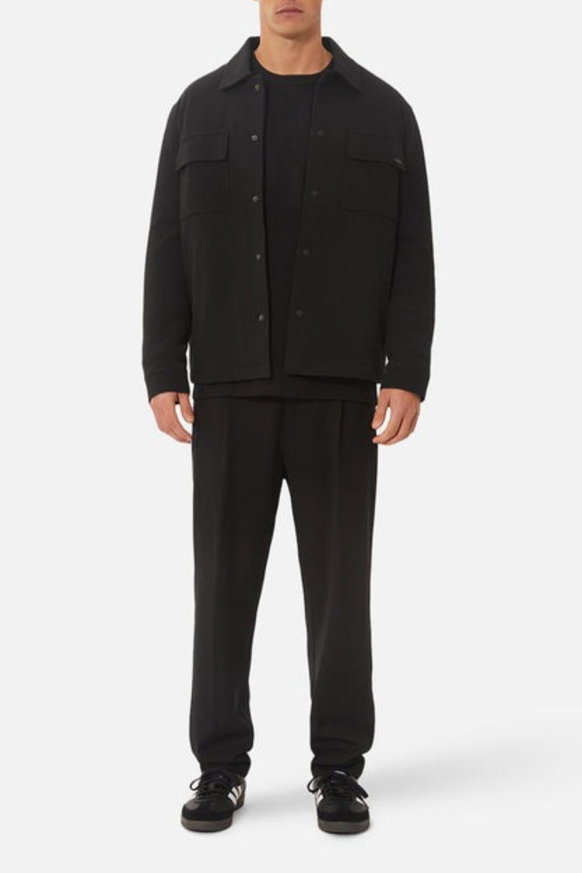 A man faces forward against a white background wearing the Industrie The Westport Jacket in black, featuring twin chest pockets, along with a black shirt, black pants, and black sneakers with white soles.
