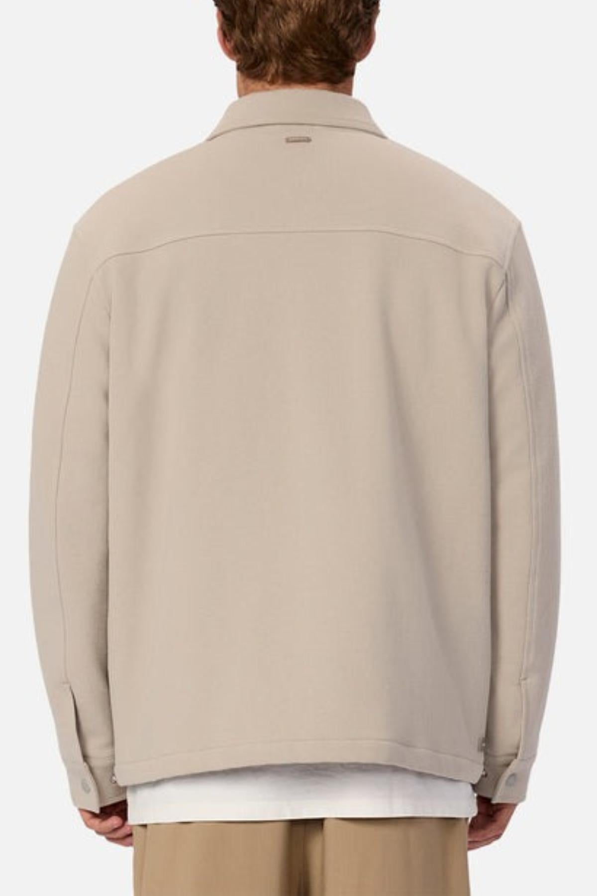 A person is seen from behind wearing the Industrie The Westport Jacket in light beige, featuring twin chest pockets, over a white shirt and beige pants against a plain, light-colored background.