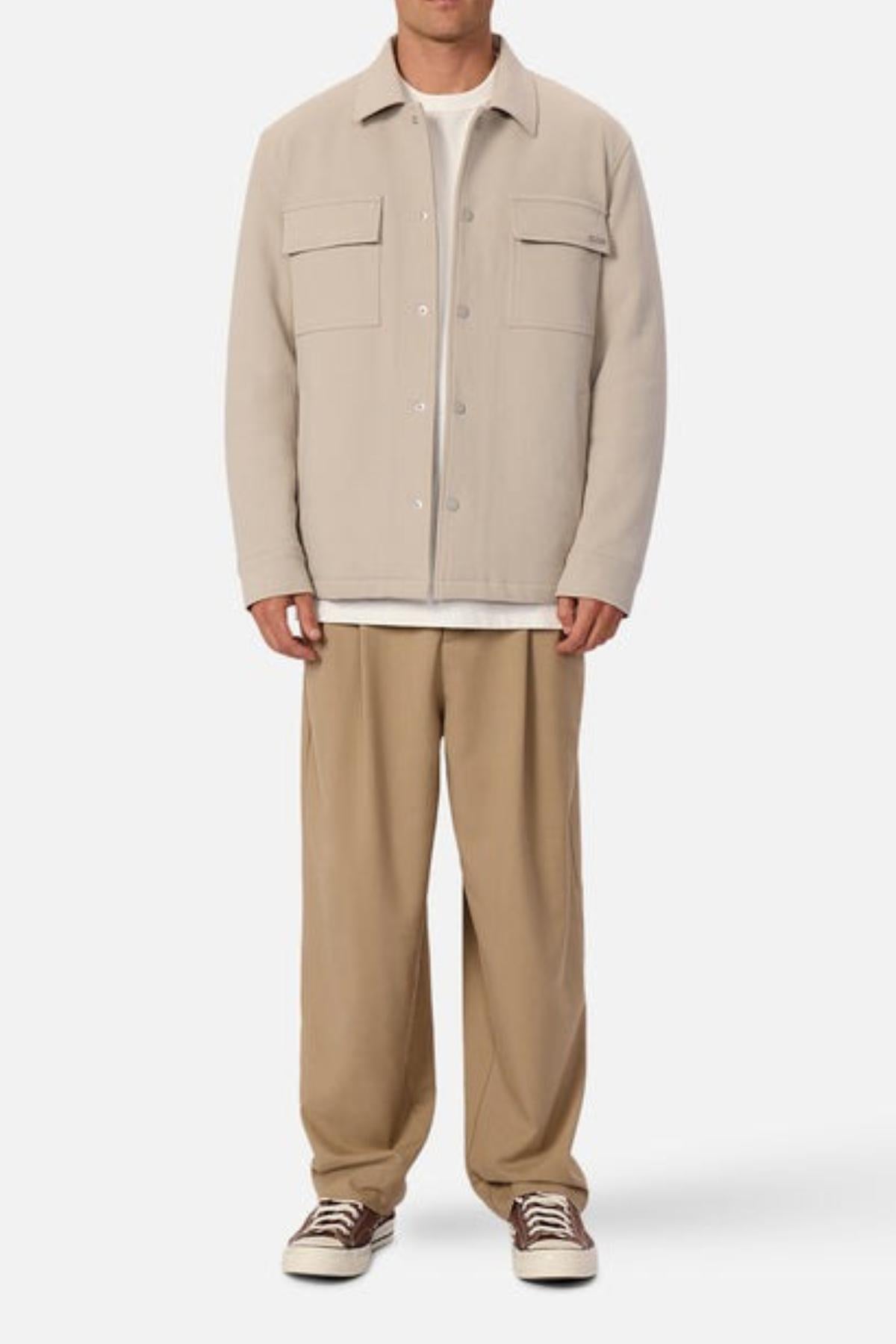 A man faces forward against a white background, wearing an Industrie The Westport Jacket in light beige with twin chest pockets over a white shirt, loose tan pants, and beige low-top sneakers featuring white laces.