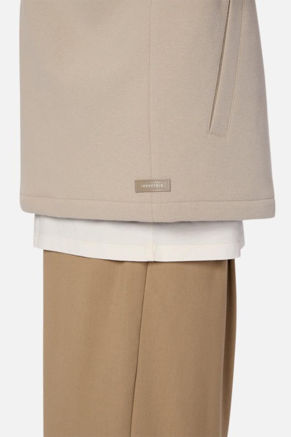 A close-up of a person wearing the Industrie The Westport Jacket, a beige coat with a minimalist design, subtle twin chest pockets, and a small rectangular label near the hem, styled over a white shirt and tan trousers.