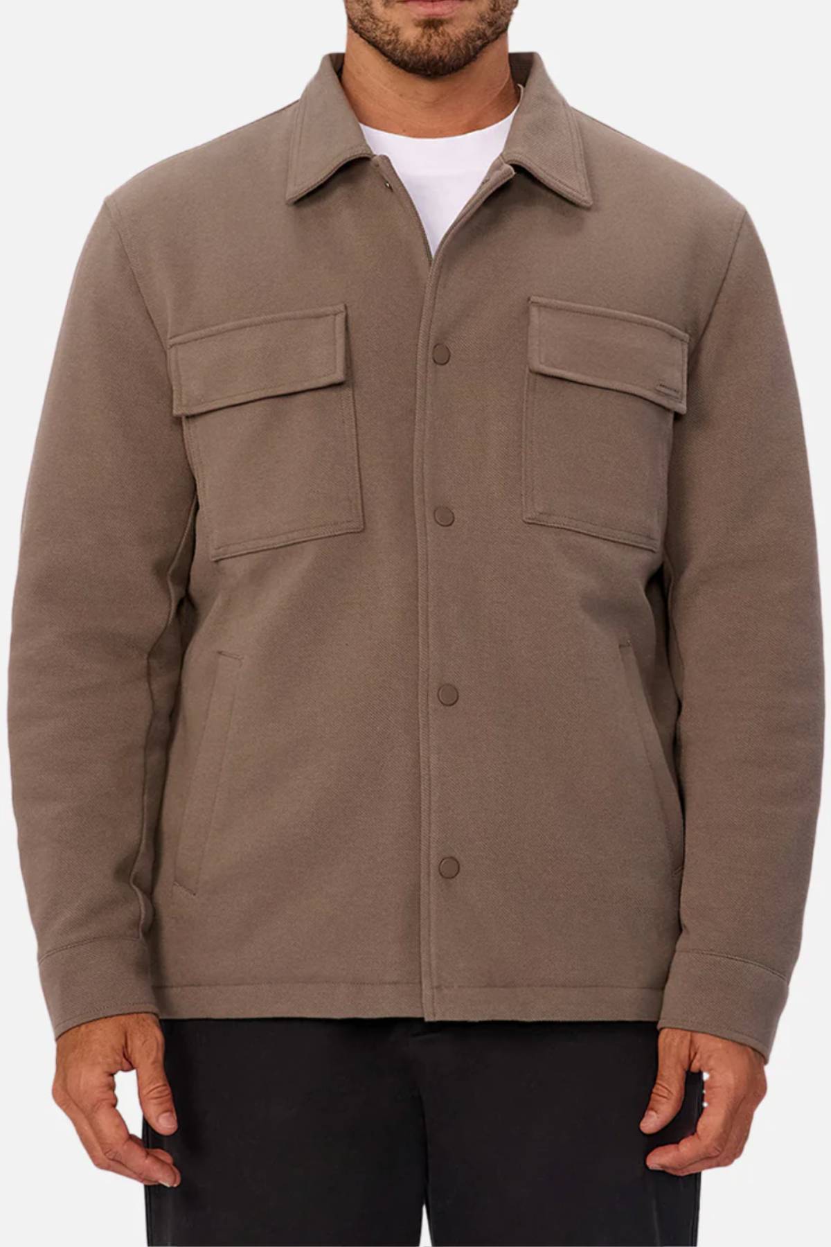 Someone wearing the Industrie The Westport Jacket—a taupe men’s jacket featuring chest flap pockets, side pockets, and a classic collar—over a white shirt and black pants; an ideal choice for everyday style.
