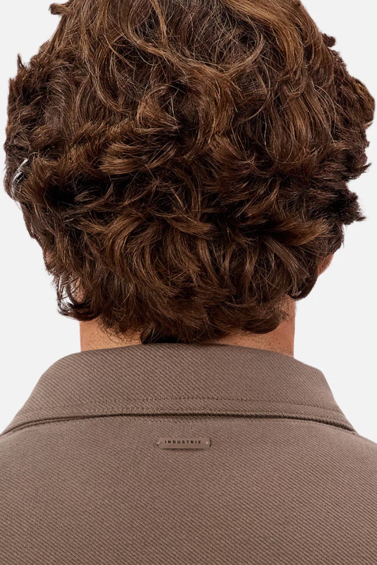 A person with wavy brown hair is seen from behind, wearing the Industrie The Westport Jacket in taupe with a small "INDUSTRIE" label on the upper back—ideal for everyday wear or layering.