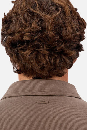 A person with wavy brown hair is seen from behind, wearing the Industrie The Westport Jacket in taupe with a small "INDUSTRIE" label on the upper back—ideal for everyday wear or layering.