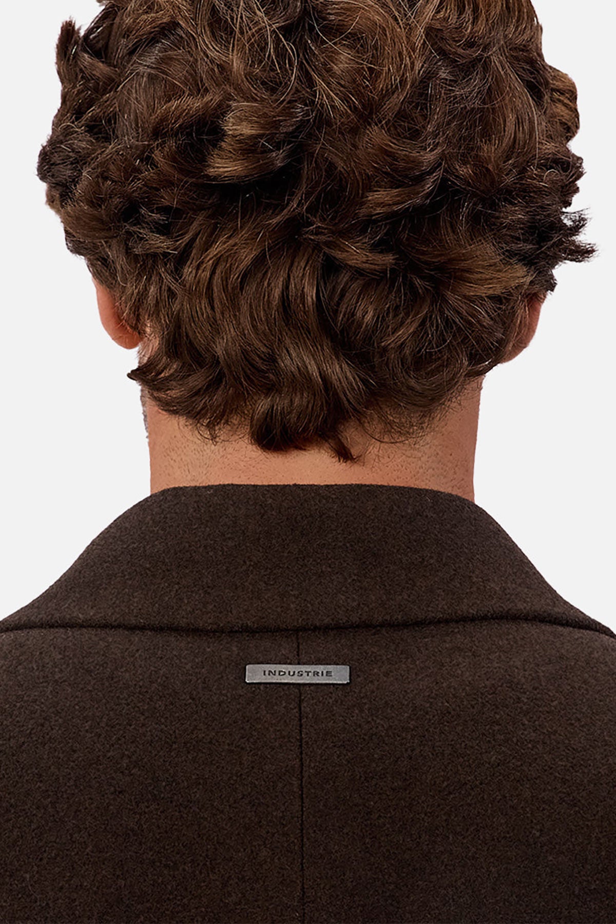 A person with wavy brown hair is shown from behind, wearing the Industrie The Wool Blend Jacques Coat in dark brown with a small metallic "INDUSTRIE" label on the back collar. The background is plain and light-colored.