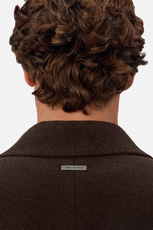 A person with wavy brown hair is shown from behind, wearing the Industrie The Wool Blend Jacques Coat in dark brown with a small metallic "INDUSTRIE" label on the back collar. The background is plain and light-colored.