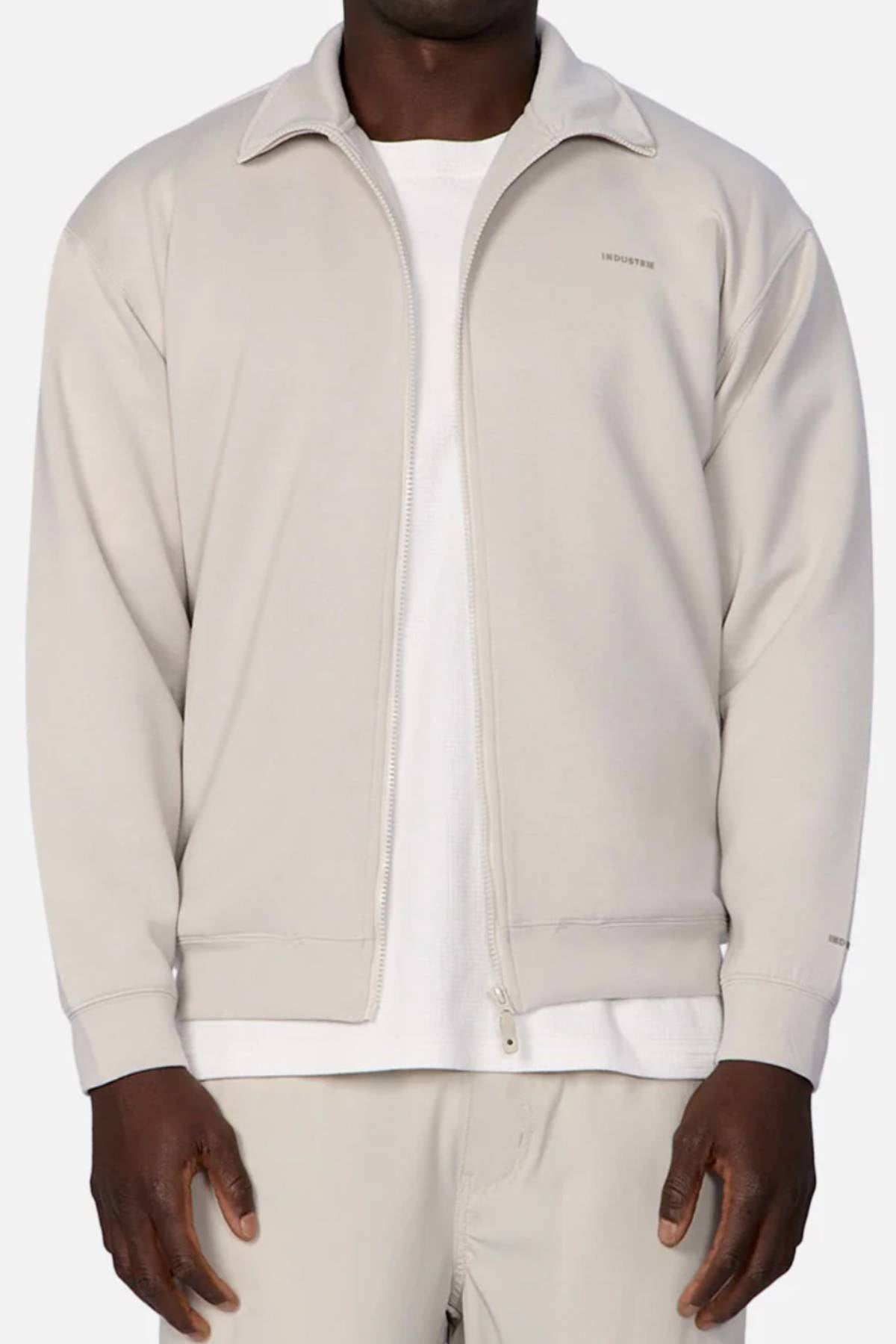 A person wears the Industrie The Zip Thru Track Top in light beige, featuring a collar and subtle left chest branding, layered over a white tee with matching beige pants, standing against a plain background.