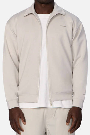 A person wears the Industrie The Zip Thru Track Top in light beige, featuring a collar and subtle left chest branding, layered over a white tee with matching beige pants, standing against a plain background.