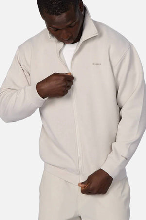 A man wearing the Industrie The Zip Thru Track Top over a white shirt looks down as he pulls up the zipper; matching beige pants complete his athletic-inspired outfit.
