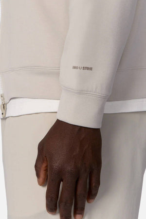 A person wearing light beige Industrie The Zip Thru Track Top, with left hand and wrist visible. The sleeve displays subtle INDUSTRIE branding, and the image is cropped to highlight the torso and stylish track top details.