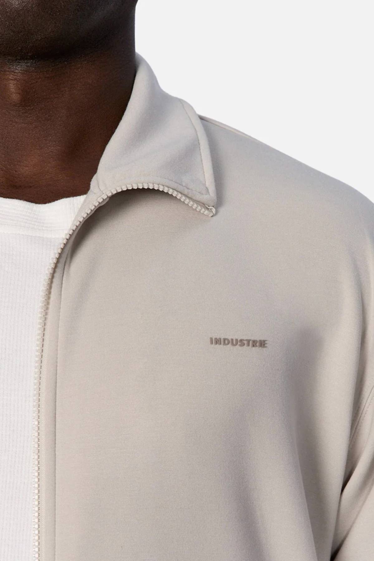 Close-up of a person wearing the Industrie The Zip Thru Track Top in light beige, featuring bold INDUSTRIE chest embroidery, layered over a white ribbed shirt. Only the lower face, neck, and upper chest are visible.