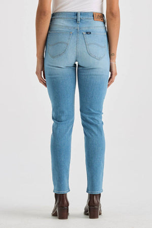 A person wearing Lee Originals LMW Mid Rise Slim Jeans in light blue and brown ankle boots, seen from the back. The jeans have back pockets and a "Lee" patch on the waistband. They stand against a plain white background.