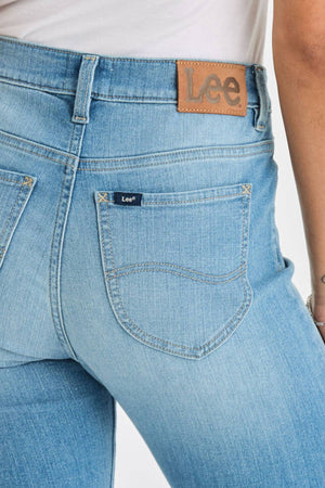 Close-up of a person wearing light blue Lee Originals LMW Mid Rise Slim Jean and a white top, showing the back pockets and brown leather Lee patch on the waistband.