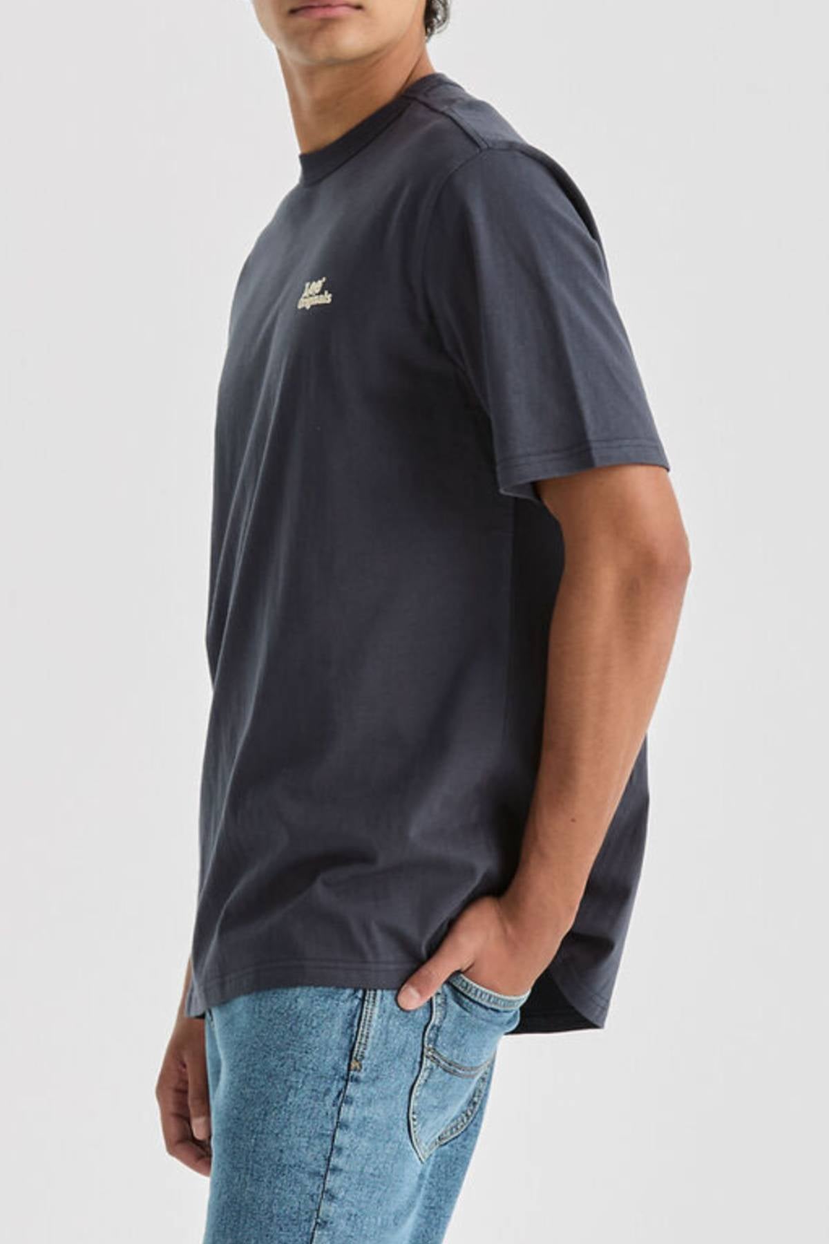Wearing a dark Lee Originals Relaxed Tee crafted from 100% cotton with a small chest logo, paired with light blue jeans, a person stands against a plain light background, turned slightly to the side with one hand in their pocket.