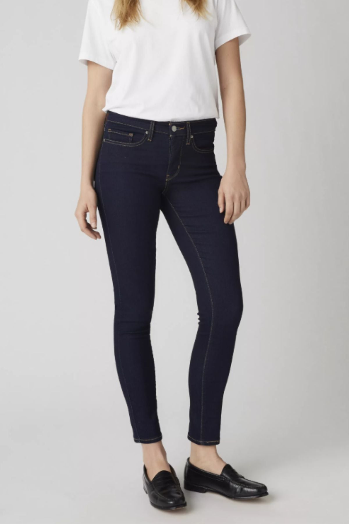 Person wearing a plain white t-shirt, Levis 311 Shaping Skinny Jean in dark blue, and black loafers, standing against a light gray background.