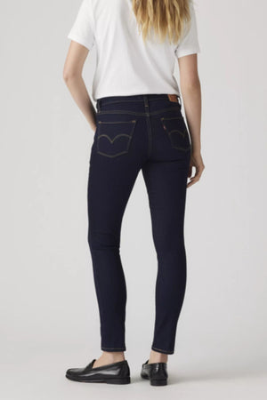 A person stands facing away, wearing a white t-shirt, black loafers, and dark blue Levis 311 Shaping Skinny Jeans made from stretch denim against a plain light background.