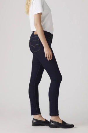 A woman stands in profile wearing a white t-shirt, black loafers, and Levis 311 Shaping Skinny Jeans made from stretch denim. She has loose blonde hair and rests her arms at her sides against a plain light background.