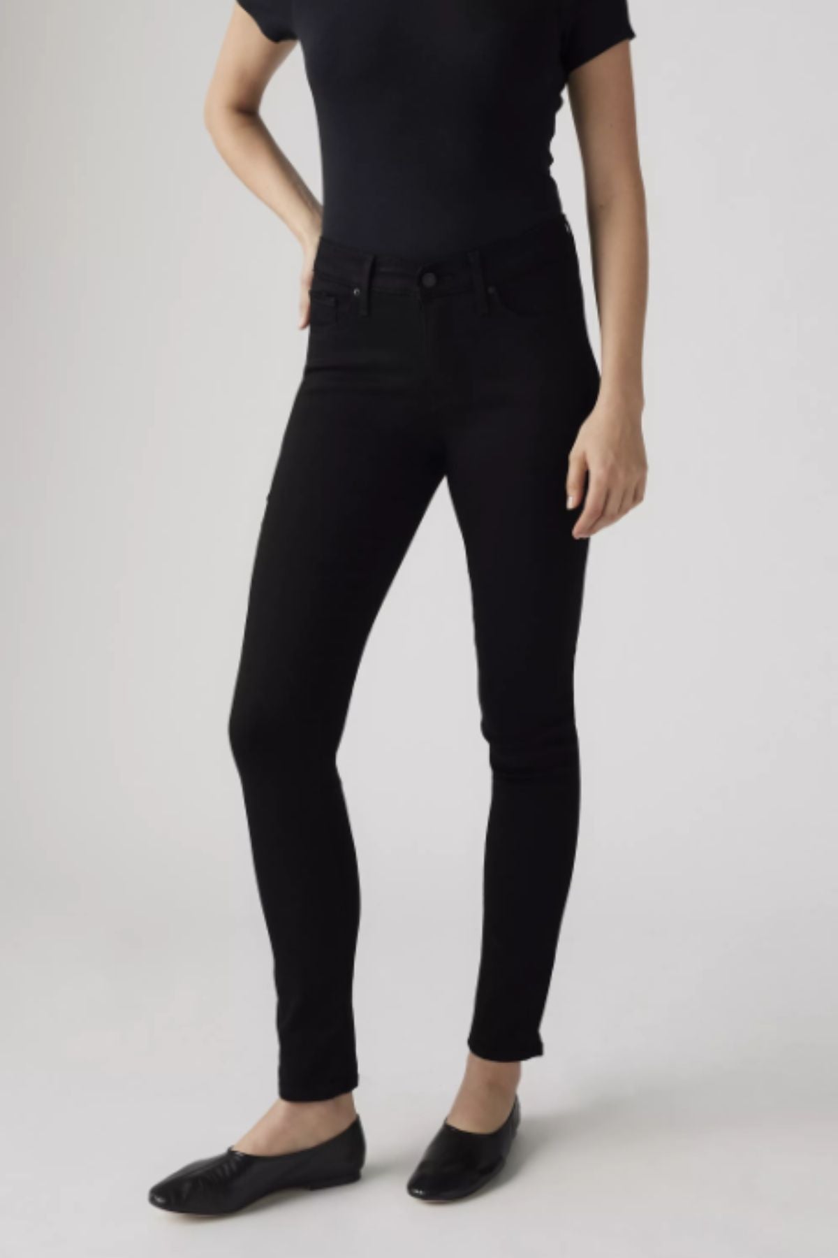 A person stands against a plain background, wearing a fitted black short-sleeve top, Levis 311 Shaping Skinny Jean in black, and black flat shoes. Their face is not visible.