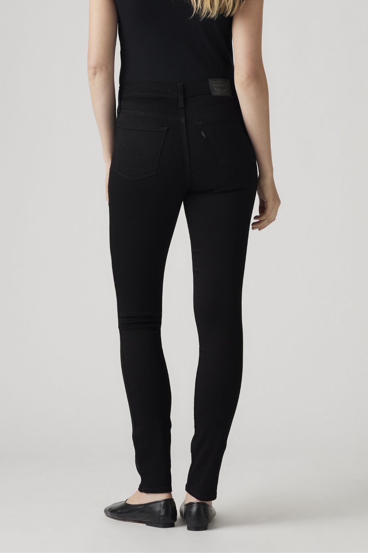 A person with long blonde hair, in a black top, Levis 311 Shaping Skinny Jean, and black flats stands with their back to the camera against a plain light gray background.