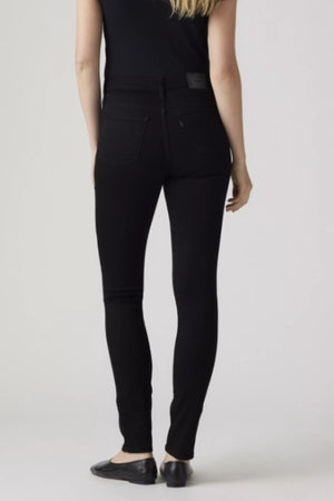 A woman stands with her back to the camera, wearing a sleeveless black top, Levis 311 Shaping Skinny Jean in black stretch denim, and black flats against a plain light background.