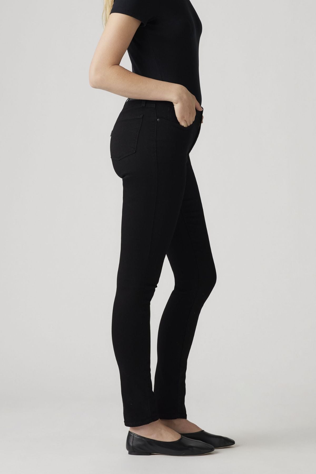 A person stands sideways against a light gray background, wearing a fitted black t-shirt, Levis 311 Shaping Skinny Jean, and black flats. Their left hand is in their pocket and their face is not visible.