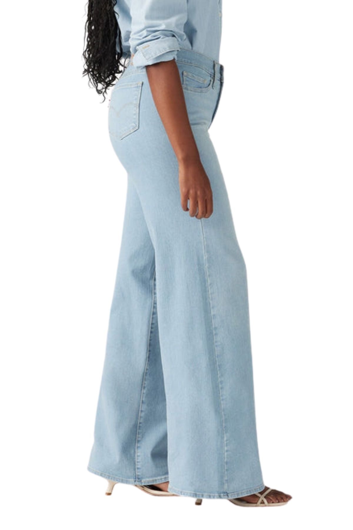 A person wearing Levis 318 Shaping Wide Leg Jean in light blue, a matching denim shirt with rolled-up sleeves, and beige heeled sandals, viewed from the side against a white background.