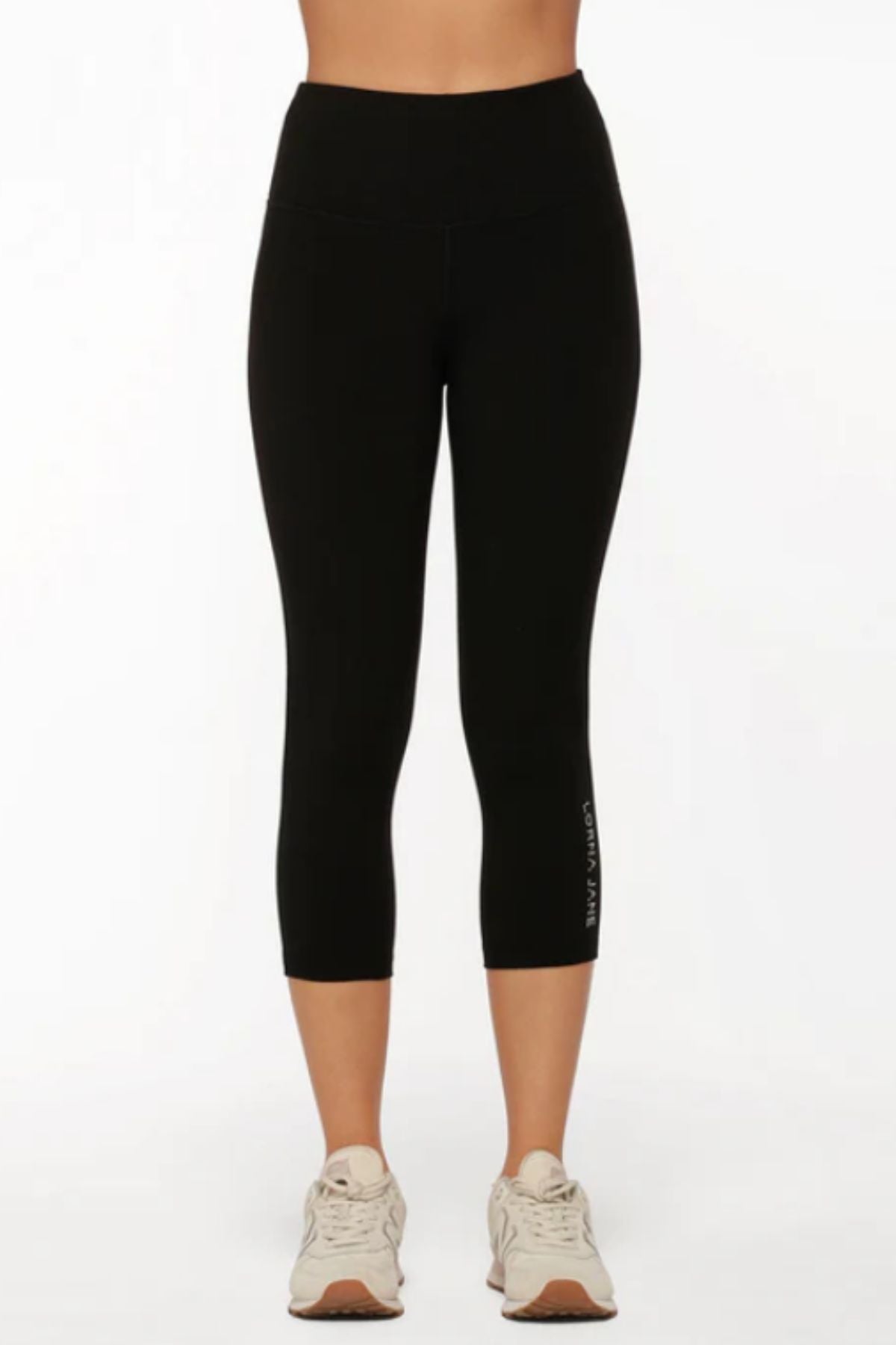 A person wears black, high-waisted Lorna Jane Lotus No Chafe Phone Pocket 7/8 Leggings and beige sneakers, standing facing forward against a plain white background for a stylish, athletic look.