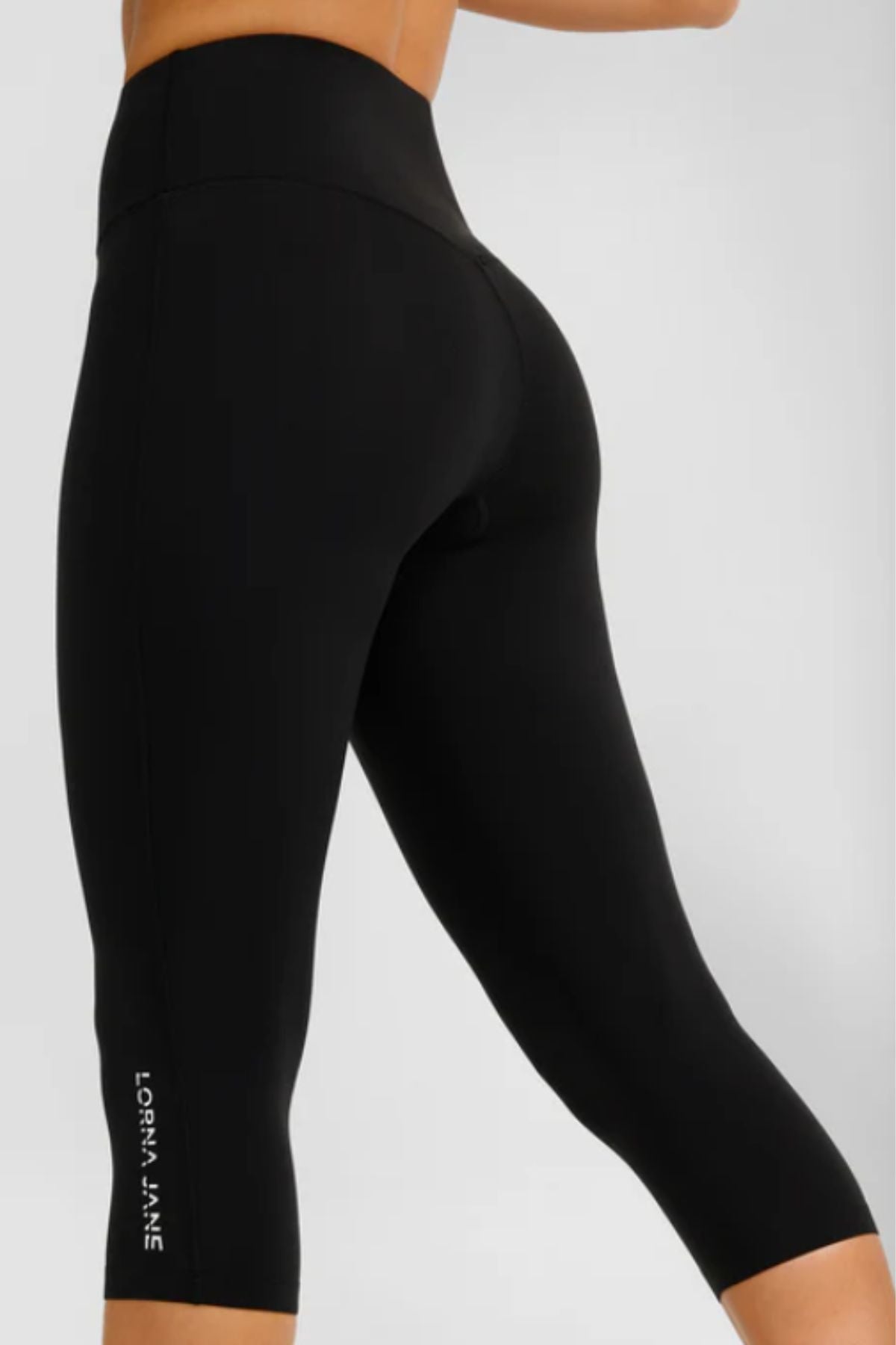 A person wears Lorna Jane Lotus No Chafe Phone Pocket 7/8 Leggings in black, featuring the white Lorna Jane logo near the lower leg. The photo, taken from the back, highlights the flattering fit of these high-waisted workout leggings.