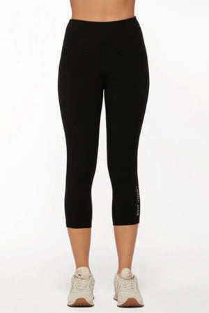 A person wears black, high-waisted Lorna Jane Lotus No Chafe Phone Pocket 7/8 Leggings and beige sneakers, standing facing forward against a plain white background for a stylish, athletic look.
