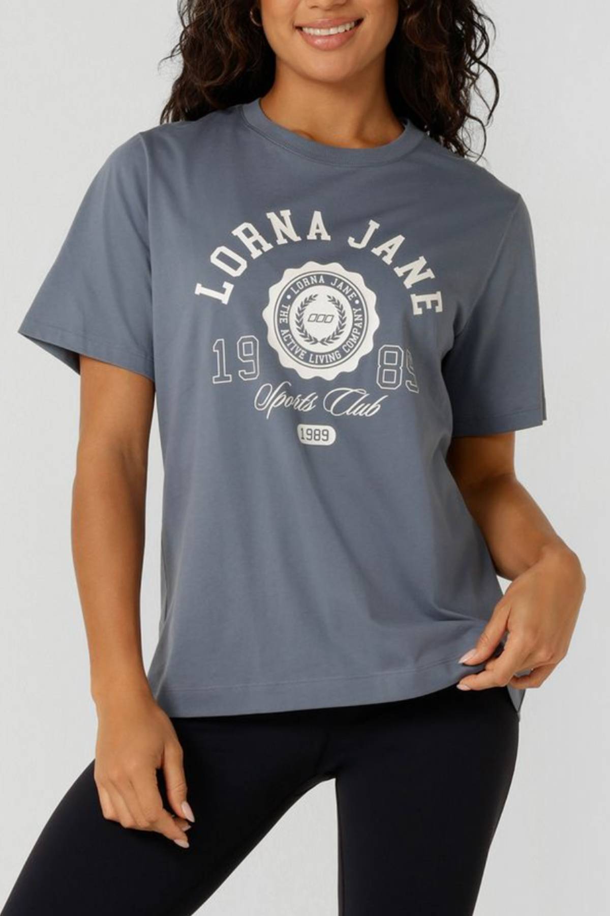 A woman smiles with one hand on her hip, wearing the Lorna Jane Active Living Relaxed T-Shirt featuring a limited edition "Sports Club 1989" logo and black leggings.