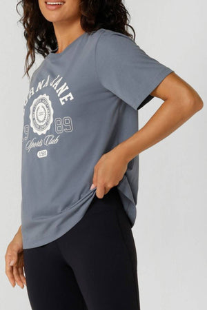 A woman wearing a loose gray Lorna Jane Active Living Relaxed T-Shirt with a limited edition logo and black leggings stands, holding the side of her shirt and smiling. Only the upper half of her body is visible.