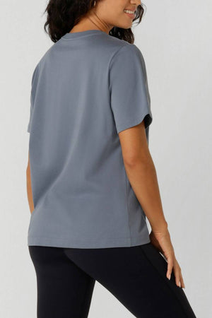 A woman with wavy dark hair wears the Lorna Jane Active Living Relaxed T-Shirt and black leggings. She smiles, facing away from the camera with her head turned slightly to the side.