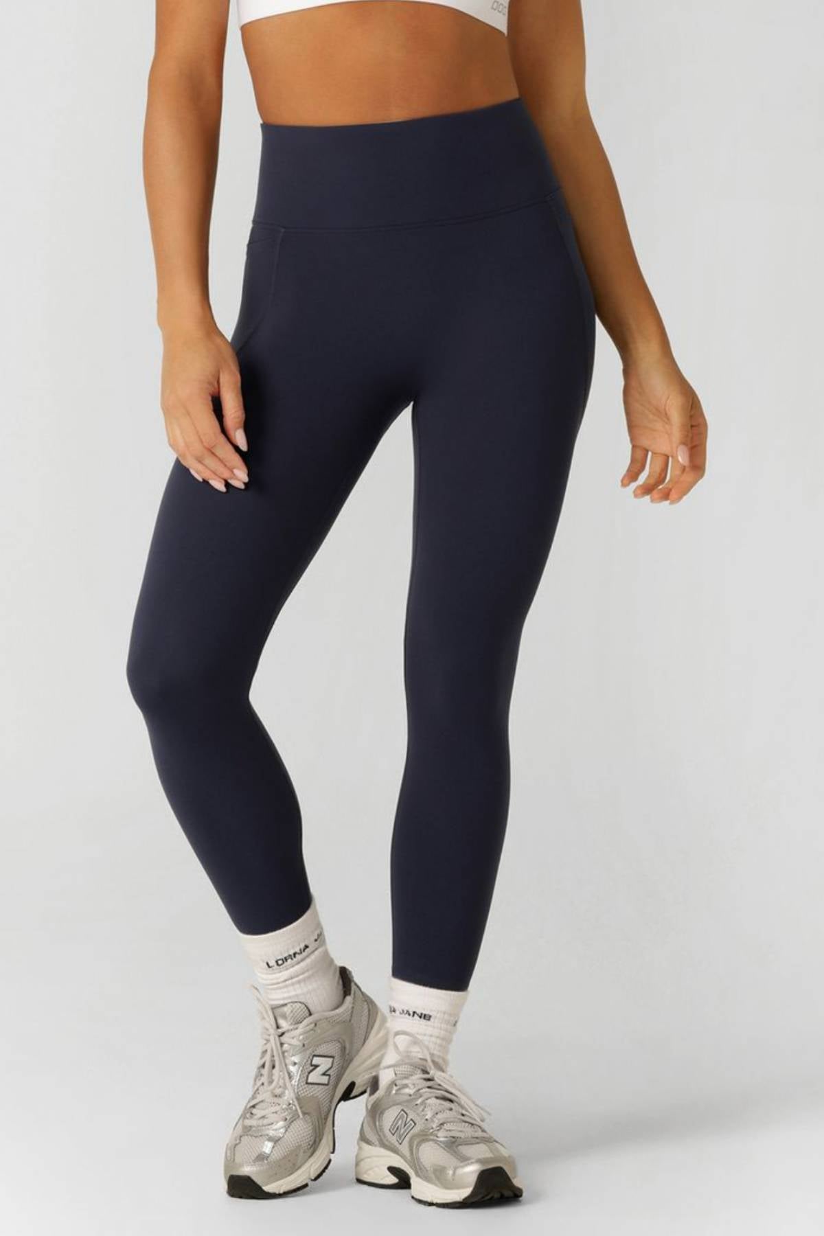 A woman wears a white sports bra, gray New Balance sneakers, white socks, and Lorna Jane Amy No Ride Phone Pocket Ankle Biter Leggings in navy blue with contouring panels. Her head is out of frame against a plain light background.