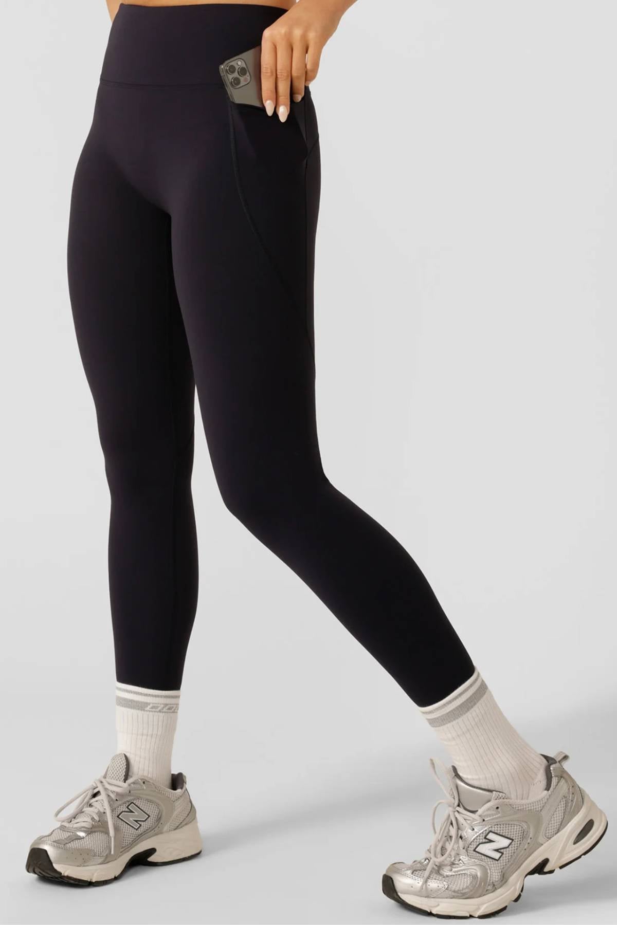 A person wearing Lorna Jane Amy No Ride Phone Pocket Ankle Biter Leggings with side phone pockets, white socks, and gray sneakers places a smartphone in the pocket. The image is cropped at the waist with a plain light gray background.