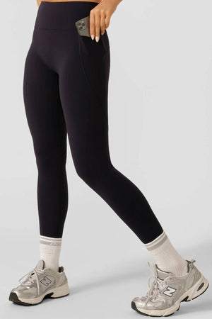 A person wearing Lorna Jane Amy No Ride Phone Pocket Ankle Biter Leggings with side phone pockets, white socks, and gray sneakers places a smartphone in the pocket. The image is cropped at the waist with a plain light gray background.