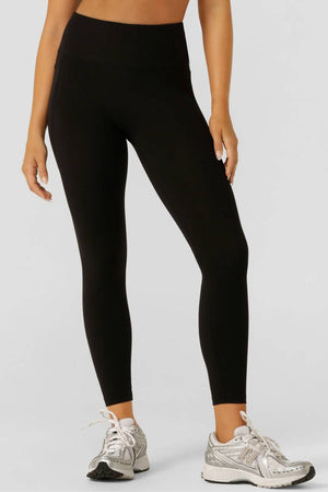 A person stands against a plain light gray background, arms relaxed at their sides, wearing black Lorna Jane Amy No Ride Phone Pocket Ankle Biter Leggings and silver athletic sneakers.