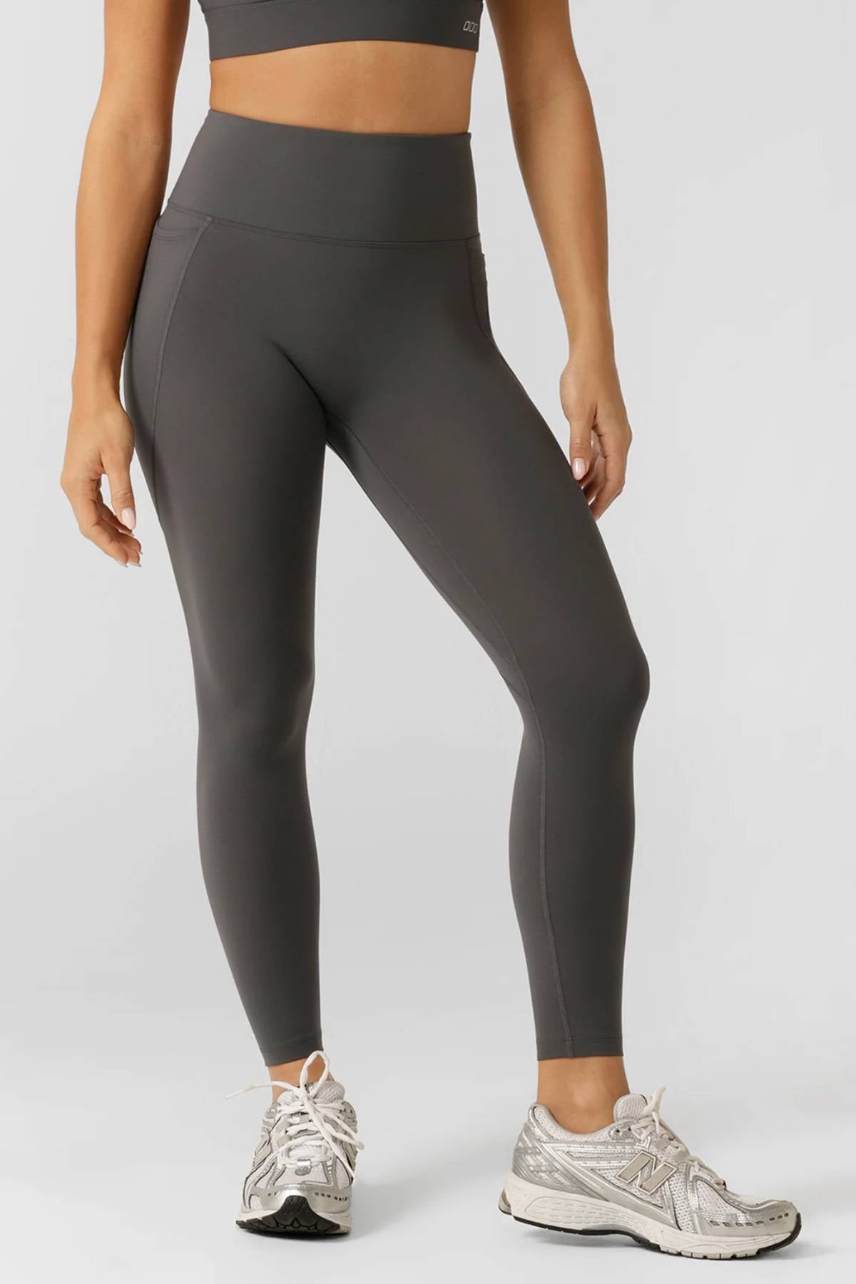 A person wears dark gray Lorna Jane Amy No Ride Phone Pocket Ankle Biter Leggings with side phone pockets and a matching sports bra, standing on a light gray background in white and gray athletic shoes. The photo is cropped below the shoulders.