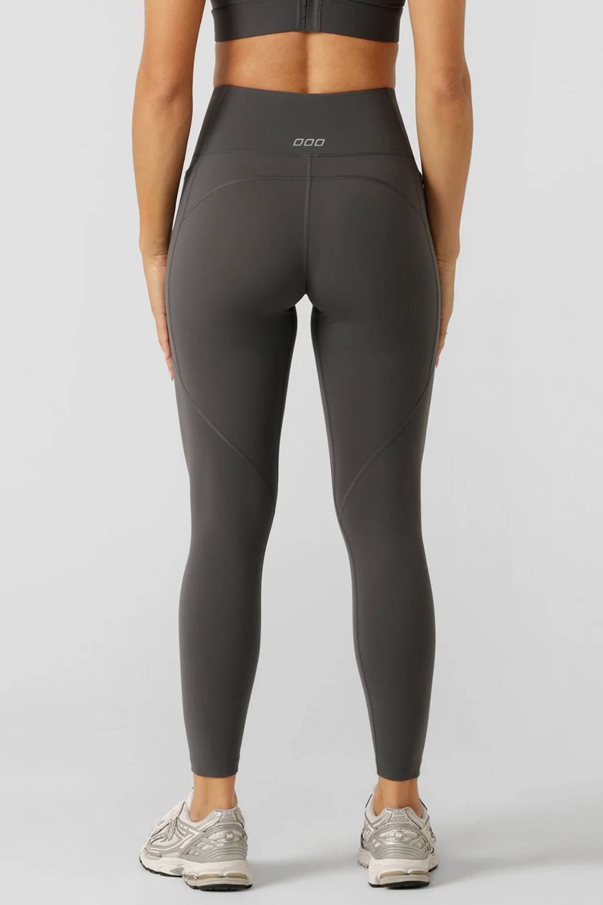 A woman models a gray sports bra and Lorna Jane Amy No Ride Phone Pocket Ankle Biter Leggings with side pockets, standing to show the back of her outfit. She pairs the look with light sneakers against a plain gray background.